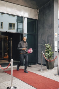 Man with bouquet of flowers using phone while walking on red carpet outside hotel