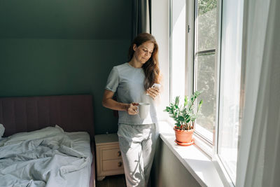 A young woman stands by the window and types on her mobile phone.