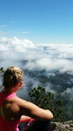 Rear view of woman sitting on cliff against cloudscape