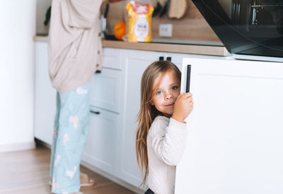 Cute little girl with long hair looks out from behind door in kitchen at home. daughter helps mother 