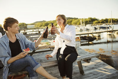 Smiling male and female friends toasting beer bottles at harbor