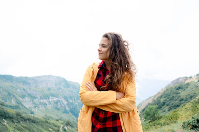 A young beautiful woman with long curly hair stands against the backdrop of a mountain landscape.