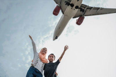 Low angle view of siblings with hand raised standing against airplane flying in sky