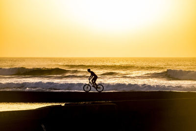 Silhouette man riding bicycle on shore at beach against clear sky during sunset