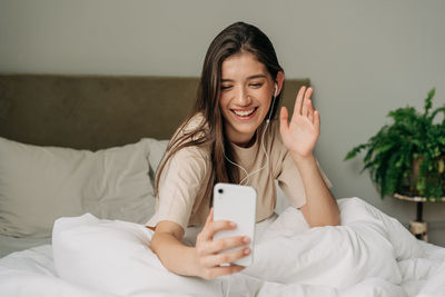 Woman sitting on bed after waking up and talking on video call using mobile phone and headphones