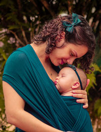 Mother carrying toddler daughter on baby sling against tree