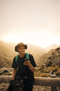 Smiling young man using mobile phone while sitting on railing against sky