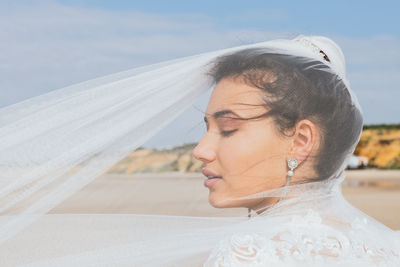 Back view of gentle pensive woman in bridal wear with lace ornament and flying veil with eyes closed on coast