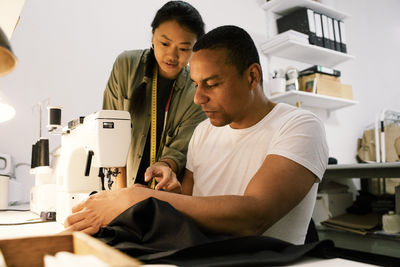 Female colleague assisting male tailor stitching clothes using sewing machine at workshop