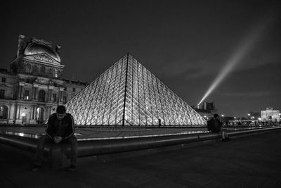 Rear view of man sitting against illuminated city at night
