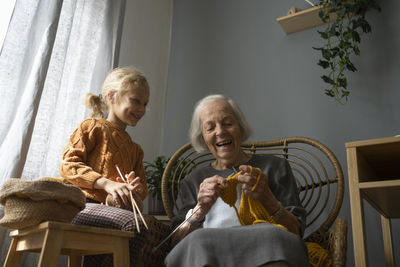 Happy girl holding ball of wool and needle looking at grandmother knitting on chair