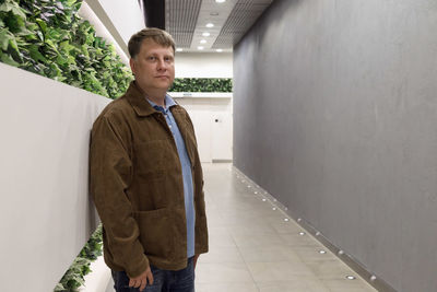 An adult tall man in a corduroy brown jacket shopping center stands against a wall with greenery.
