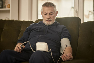 Senior man checking blood pressure sitting on sofa at home