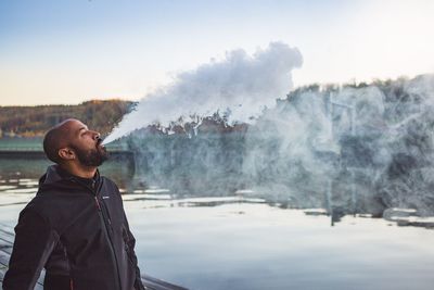 Side view of mid adult man emitting smoke while standing by lake against sky
