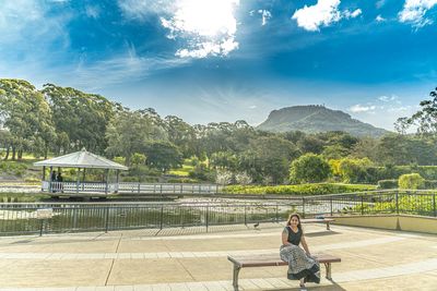 Woman sitting on bench by trees against sky