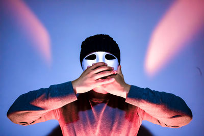 Close-up of man covering mouth while wearing mask against illuminated wall