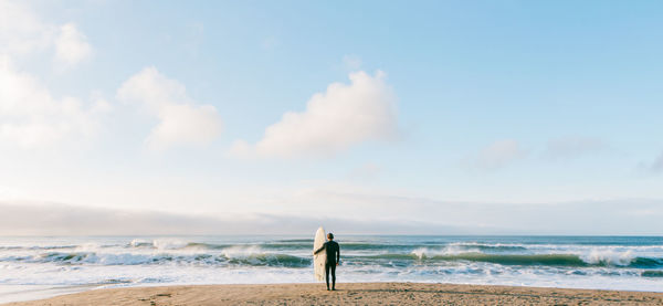 Rear view of man with surfboard standing at beach against sky