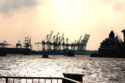 Ship moored on river by cranes against sky during sunset