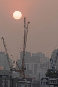 Cranes by buildings against sky during sunset