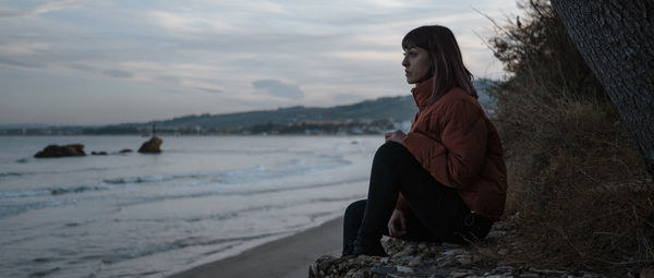 Woman sitting on rock at beach against sky