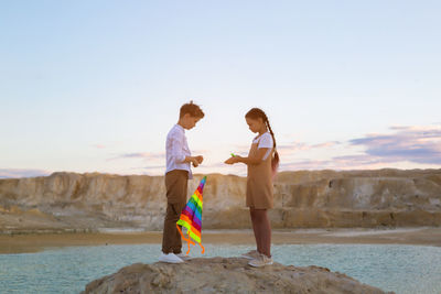 A boy and a girl are standing with a kite on a high stone near the lake.