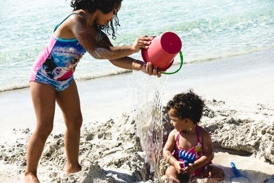 Side view of girl pouring water on sister sitting at beach