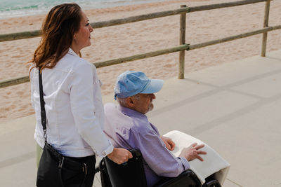 Delighted adult daughter pushing wheelchair with senior father and enjoying stroll along promenade near sea