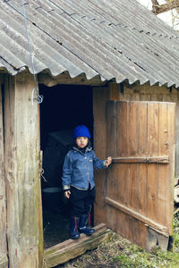 Village boy in boots stands at the door of the barn in warm clothes