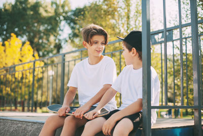 Two cute teenagers sit in a skatepark, relax after skateboarding and chat. 