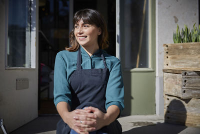 Smiling owner looking away while sitting outside store during sunny day