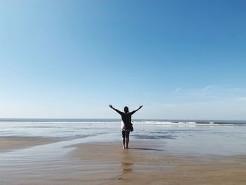 Rear view of woman with arms raised standing on beach against blue sky