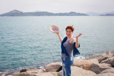 Portrait of cheerful woman standing on rocky shore against cloudy sky