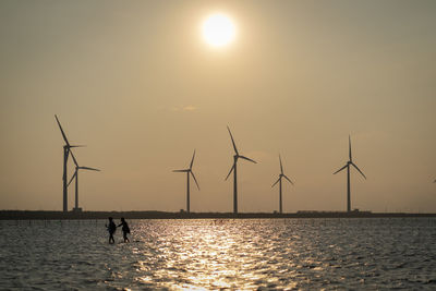 Silhouette wind turbines in sea against sky during sunset