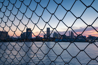 City seen through chainlink fence against sky during sunset