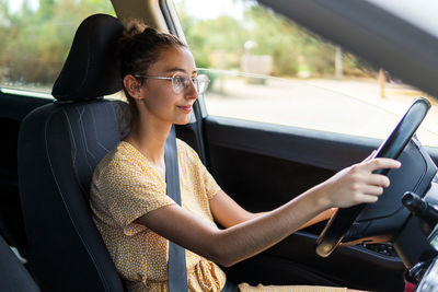 Young female in casual clothes and glasses looking away and driving contemporary vehicle on sunny day