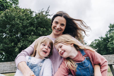 Cheerful mother with arms around daughters sitting on bench