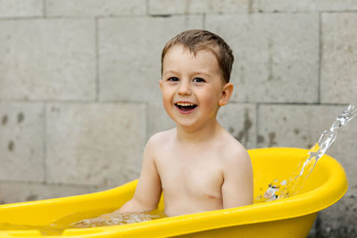 Cute little boy bathing in yellow tub outdoors. happy child is splashing, playing with water 