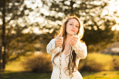 Portrait of beautiful woman holding chain standing against trees at park