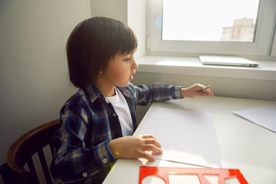 Boy child draws on paper with a ruler on a table sitting by the window