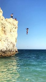 Man jumping on rock in sea against clear blue sky