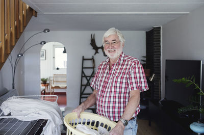 Portrait of smiling senior man holding laundry basket while standing in living room