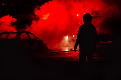 Rear view of silhouette man standing by smoke on illuminated street at night