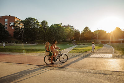 Teenage girl sitting with friend riding bicycle at park during sunset