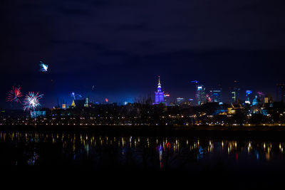 Illuminated buildings by river against sky at night