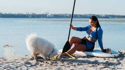 Young woman sitting on sup board and laughing at her japanese spitz digging a hole on the beach