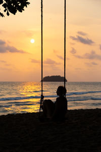 Silhouette woman sitting on swing at beach against sky during sunset
