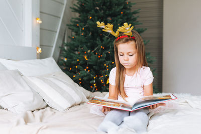 Cute little girl in pink dress reading book sitting on bed in room with christmas tree