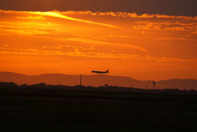 Airplane flying over silhouette field against sky during sunset