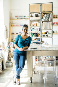 Full length portrait of confident businesswoman leaning on table in office