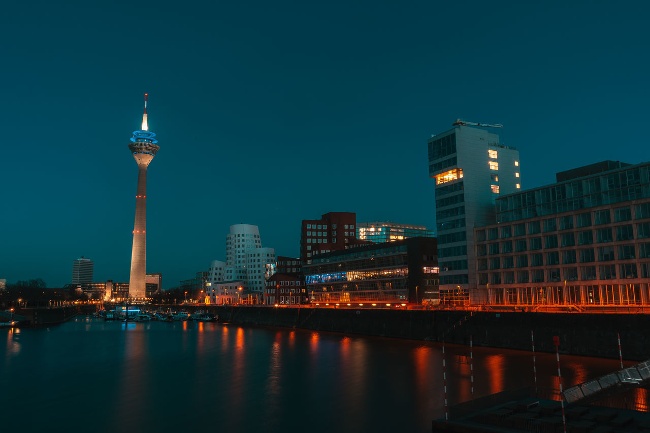 ILLUMINATED MODERN BUILDINGS BY RIVER AGAINST SKY AT NIGHT